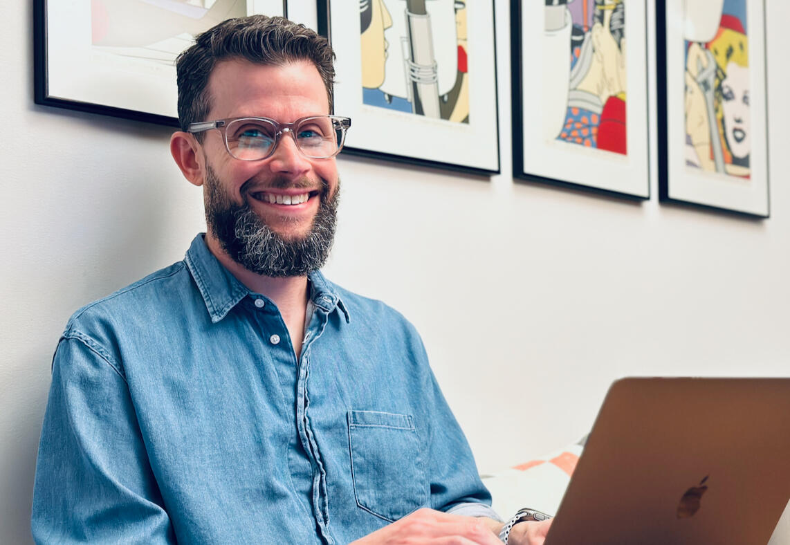 A casual photo of a man, smiling at the camera, sitting at his computer with artwork on the wall behind him.